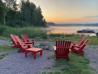 A circle of red Adirondack chairs surrounds a fire pit near a tranquil lake at dawn. Concept of: Lakeside relaxation.