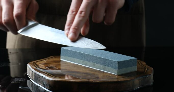 Man sharpening knife with sharpener at mirror table against black background, closeup