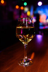 Glass of white wine on a wooden bar counter with colorful bokeh lights in the background. Cozy nightlife atmosphere in a modern nightclub or lounge bar