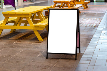 A blank white A-frame sidewalk sign stands on a tiled floor in front of some picnic tables inside...