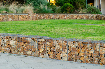 A stone-filled gabion wall used as a functional and aesthetic edge divider between a paved walkway and grass lawn. Natural textures with clean lines to create separation in an urban public space
