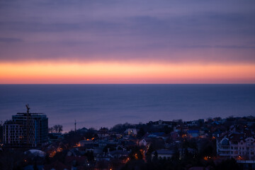 Coastal town at dusk with a vivid gradient sky transitioning from orange to purple. Calm sea reflects the light, buildings glow gently, and a crane marks urban growth on the horizon.