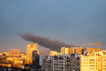 Cityscape with residential buildings and construction cranes in the morning light, with a large dark plume of smoke from a fire in the background. Urban environment and environmental pollution concept