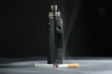 Electronic smoking device and broken cigarette in smoke on dark table against black background,...