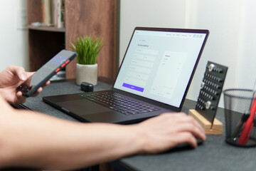 A man completing online payment method on laptop using smartphone at home office desk