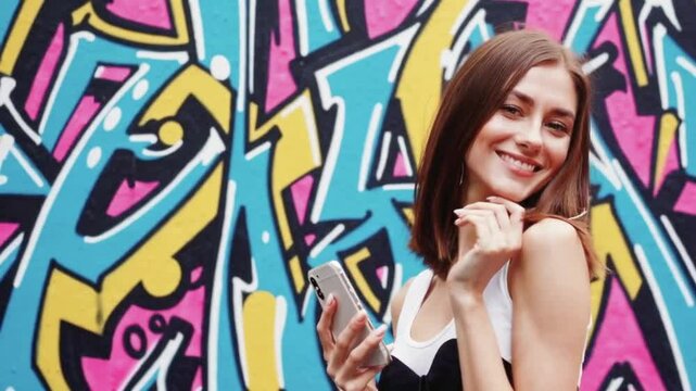 Trendy girl smiling with smartphone against graffiti wall