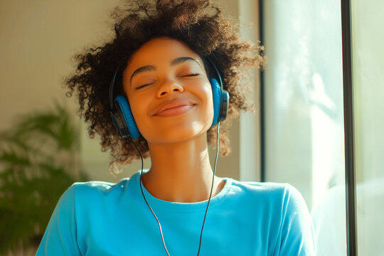Satisfied curly young Afro American woman has carefree happy mood, closes eyes and listens music in headphones, wears casual blue t shirt, poses indoor. People, leisue, entertainment concep