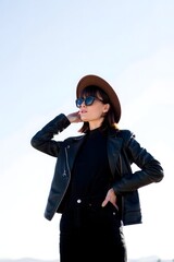 A stylish woman posing confidently in the sun, wearing a hat, sunglasses, and a leather jacket. She exudes elegance and a carefree attitude