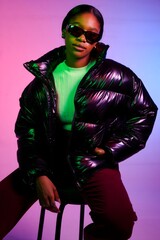 Stylish woman posing in a shiny black puffer jacket. She's sitting on a stool against a vibrant pink and blue background, wearing sunglasses. 