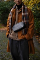 A stylish man is wearing a fashionable outfit outdoors on a cool day. The man is accessorized with a scarf and crossbody bag