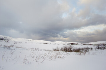 Snow-covered landscape with gentle hills, sparse vegetation, and dramatic clouds creating a serene...