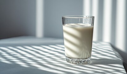 A glass of milk sits on a white surface, illuminated by sunlight streaming through blinds, casting dramatic shadows