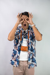 Happy young asian man wearing blue summer shirt is making peering through binoculars hand gesture,...