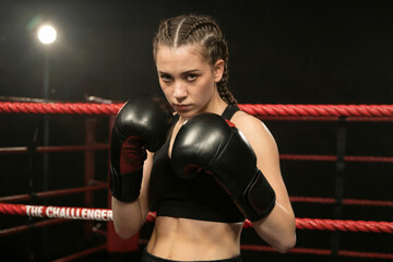 A young woman in a boxing ring, poised in a defensive stance. She wears black boxing gloves and a black sports bra. 