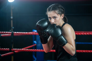 A young woman in a boxing ring, poised in a defensive stance. She wears black boxing gloves and a black sleeveless top. 