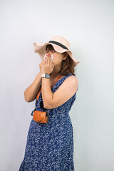 Beautiful young asian woman wearing blue summer dress and beach straw hat is sneezing and covering using tissue paper, isolated over white background.