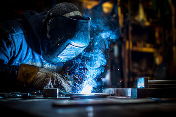 A skilled industrial welder wearing safety gear, including gloves and a protective helmet with visor, working in a dimly lit metal workshop