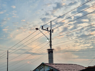 Electric power pole with glowing streetlamp and intersecting wires under cloudy twilight sky with...
