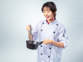 Asian female chef in white uniform holding a black saucepan and metal whisk, smiling...
