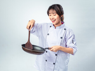 Asian female chef smiling while stirring an empty frying pan using a wooden spatula, wearing a...