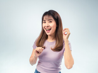 Asian woman in a light purple t-shirt smiling and raising her fists excitedly, expressing joy and...