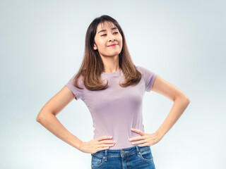 Confident Asian woman standing with hands on hips, smiling with pride and satisfaction, wearing a purple t-shirt and jeans against a clean studio background