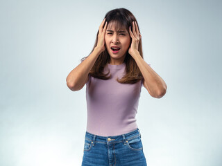 Asian woman in purple t-shirt and jeans holding her head with both hands, eyes closed, expressing...
