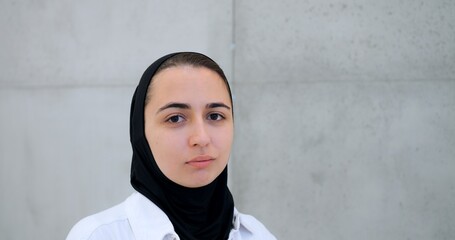 Serious young muslim woman wearing black hijab, white shirt, standing near gray concrete wall, gazing directly at viewer with neutral expression