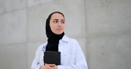 Muslim woman in hijab, white jacket holding black book, turning head sideways against concrete wall background