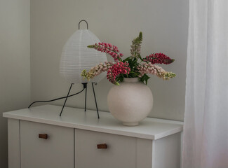 A bouquet of lupines in a ceramic vase, a scandinavian-style paper table lamp on a white chest of drawers in the living room