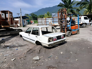 A white car, significantly damaged and disassembled at scrap yard or junkyard. The scene includes a...