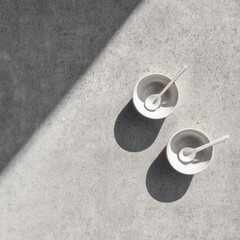 Two small white bowls with spoons, cast in sunlight on a mottled grey surface