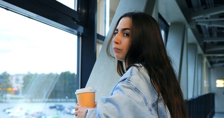 Young woman wearing a denim jacket stands in an airport terminal, holding a takeaway coffee cup and checking the time on her smartwatch while looking out the large window at the planes and runway