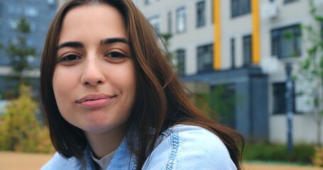 Beautiful young woman with long brown hair, wearing a light blue denim shirt, poses and looks around in front of a modern residential building