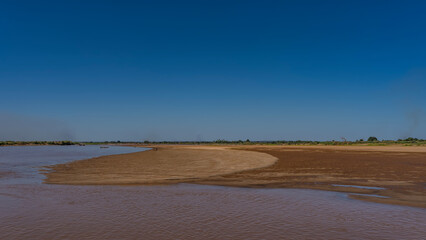 An exotic African river. Red-brown water, sandy shore. A tourist canoe floats in the distance. Green vegetation on the horizon. Clear blue sky. Copy space. Madagascar. Tsiribihina river.