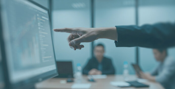 Hand pointing at a screen during a business meeting in a modern office. Data and charts are visible as colleagues work in the background with focus and intent. - Powered by Adobe