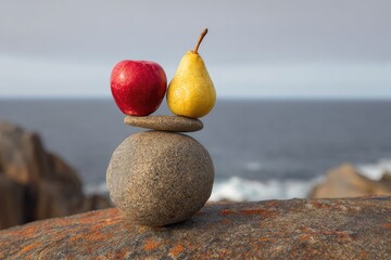 A red apple and yellow pear delicately balanced atop a three-stone cairn, set against a blurred ocean backdrop of grey-blue water and rocky coast