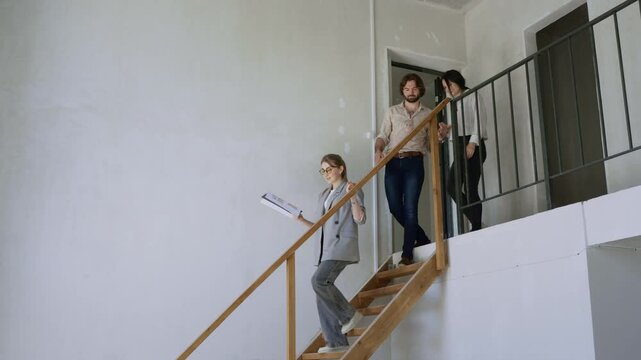 A smooth slow motion shot captures a young couple descending the stairs with their realtor, then walking through the apartment while discussing layout and blueprints on sunny day - Powered by Adobe