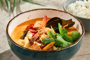 Tom yum seafood soup in rustic bright closeup, garnished herbs and chili, ceramic bowl, side of steamed rice