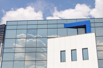 A modern office building with large windows reflecting the blue sky and white clouds, with one side...