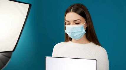 A young woman wearing a face mask points at a laptop with a blank screen against a blue background - Powered by Adobe