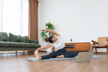 Young Asian woman stretching yoga workout on exercise mat while online training class with computer laptop in living room