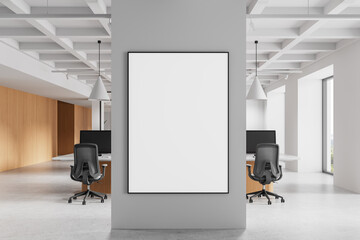 Blank poster frame on grey wall in modern open office with workstations, wood paneling, and pendant...