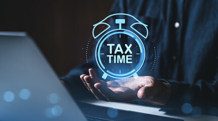 Businessman holding glowing tax time alarm clock icon over open hand with laptop in background,...