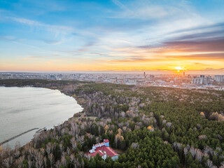 Autumn forest on lake shore at sunset and city on horizon, aerial view