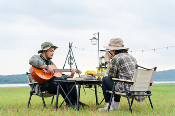 Asian couple camping in a wide meadow near mountains and a lake in the evening, relaxing with wine and music in a warm and quiet atmosphere amidst nature and the warm light of the sunset.