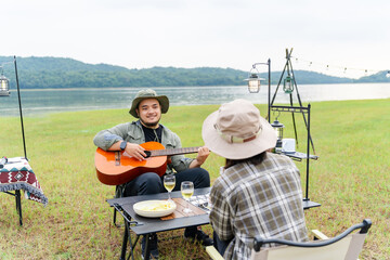 Asian couple camping in a wide meadow near mountains and a lake in the evening, relaxing with wine and music in a warm and quiet atmosphere amidst nature and the warm light of the sunset.