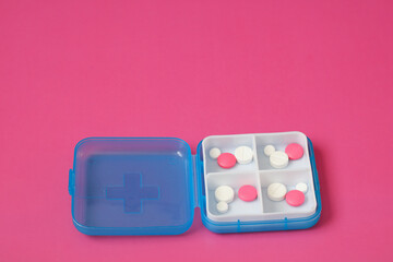 Blue pillbox containing white and pink medicine lies on pink background