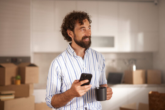A man stands in a new home holding a coffee cup and a smartphone, smiling as he enjoys a break amidst numerous unpacked boxes around him.