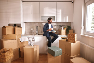 A man sits on the counter enjoying a cup of coffee amidst numerous packed boxes in a bright...